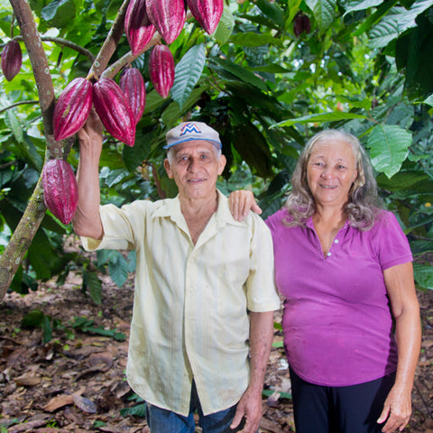 Juan Penalo and Nieve Quezada stand together in front of their cacao farm with reddish purple cacao pods growing from a cacao tree